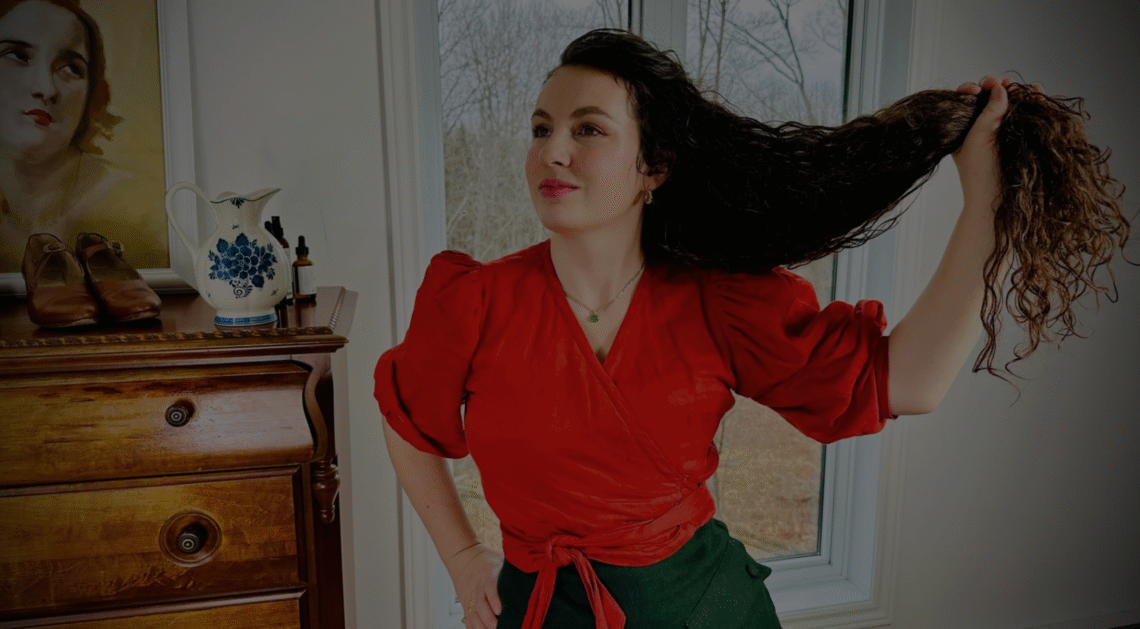 Katherine tenant ses cheveux longs dans une main. Elle porte des vêtements de style ancien.